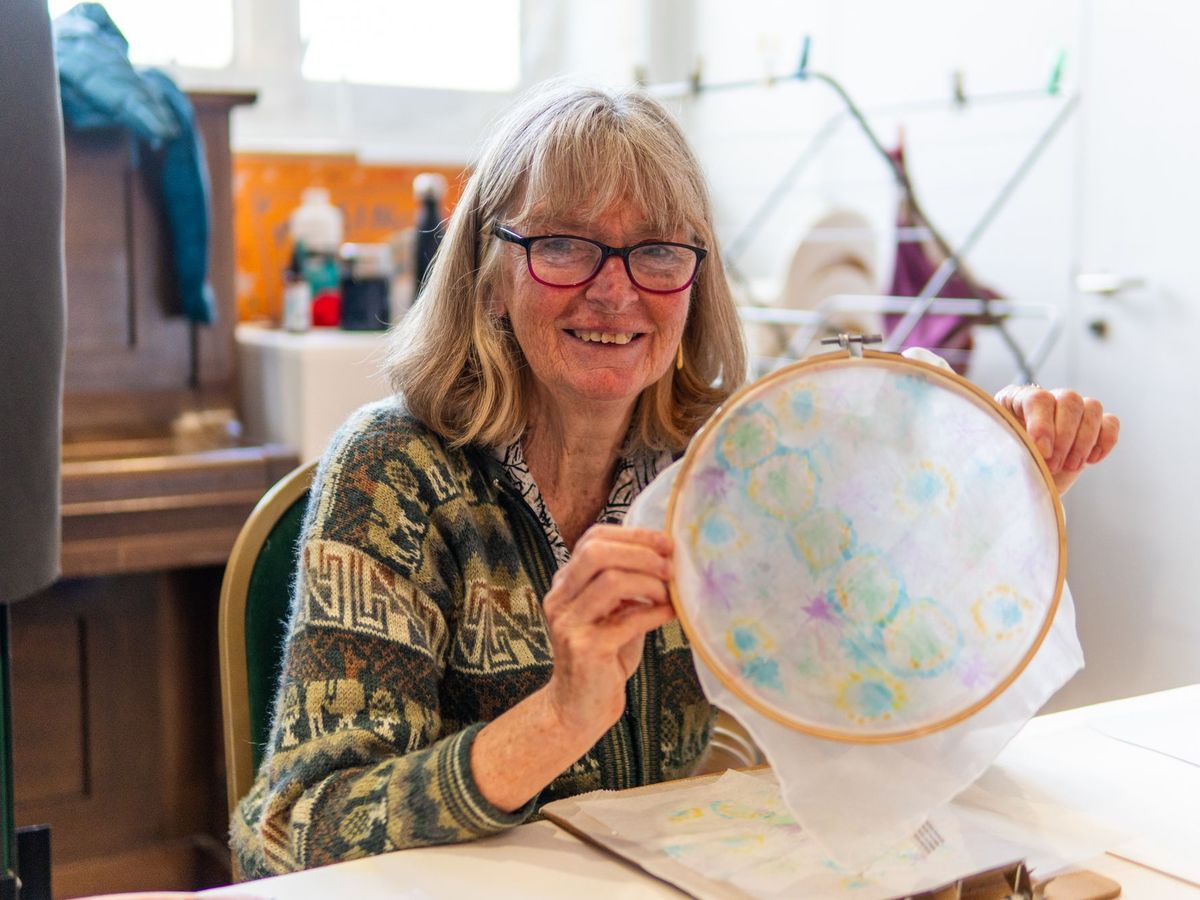 A smiling woman in glasses holds up a piece of her embroidery.