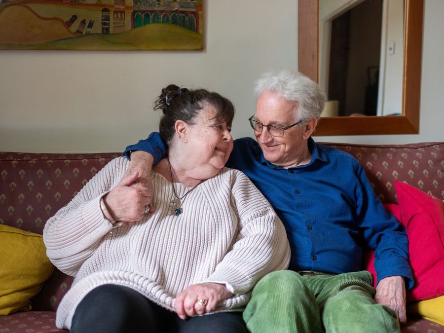 An older couple sit together on a sofa at home, smiling at each other.